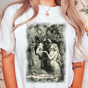 Pode incluir: Camiseta branca com uma ilustra&ccedil;&atilde;o em preto e branco de duas sereias se abra&ccedil;ando. A ilustra&ccedil;&atilde;o &eacute; cercada por uma borda preta e o texto "THE LOVERS" &eacute; impresso abaixo da imagem.