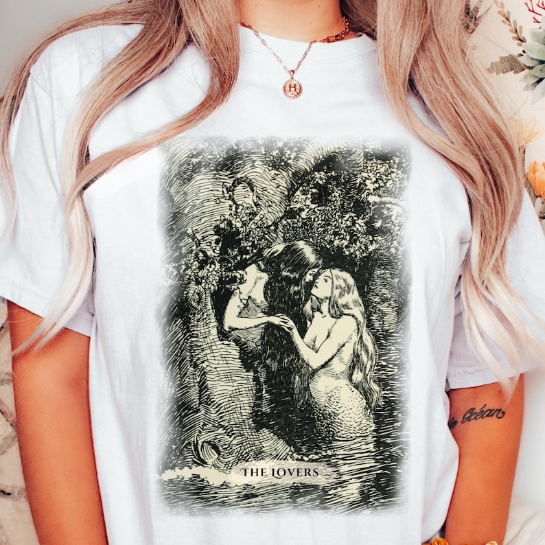 Pode incluir: Camiseta branca com uma ilustra&ccedil;&atilde;o em preto e branco de duas sereias se abra&ccedil;ando. A ilustra&ccedil;&atilde;o &eacute; cercada por uma borda preta e o texto "THE LOVERS" &eacute; impresso abaixo da imagem.