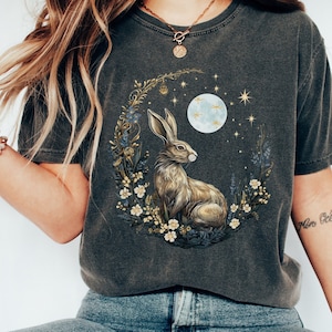 May include: A dark gray t-shirt with a graphic of a brown rabbit sitting in a crescent moon surrounded by flowers and stars. The moon is blue and white.
