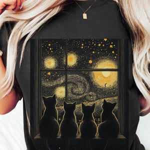 May include: A black t-shirt featuring a graphic of four black cats gazing out of a window at a starry night sky. The sky is painted in a swirling style reminiscent of Vincent van Gogh's Starry Night.