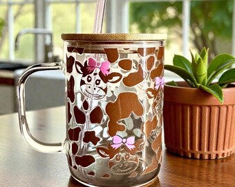 Pode incluir: Uma caneca de vidro transparente com alça, apresentando um padrão de manchas de vaca castanhas e pequenas caras de vaca com laços cor-de-rosa. A caneca inclui uma tampa de bambu e uma palhinha de vidro rosa translúcido.