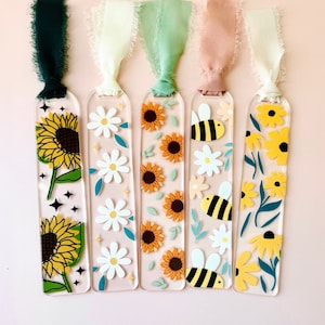 May include: Five clear acrylic bookmarks with different floral and bee designs. The bookmarks have a ribbon attached to the top. The designs include sunflowers, daisies, bees, and leaves.