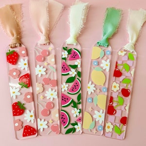 May include: Five colorful acrylic bookmarks with different fruit designs. Each bookmark features a different fruit, including strawberries, cherries, watermelon, lemons, and cherries with a checkered pattern. The bookmarks are attached to pink, white, and green ribbons.