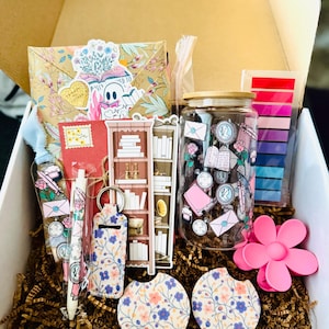 May include: A gift box filled with stationery and accessories. Includes a floral-patterned envelope, a red notebook, a pen, a keychain, a jar, two car coasters, a pink flower clip, and a bookshelf sticker. The box is filled with brown crinkle paper.
