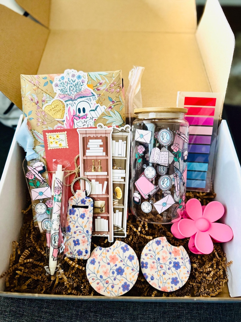 May include: A gift box filled with stationery and accessories. Includes a floral-patterned envelope, a red notebook, a pen, a keychain, a jar, two car coasters, a pink flower clip, and a bookshelf sticker. The box is filled with brown crinkle paper.