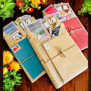 May include: A collection of handmade journals and notebooks. Each journal is wrapped in vintage newspaper and tied with twine. The journals feature colorful covers, stickers, and the text "OMANTASY" and "MEANWHILE".