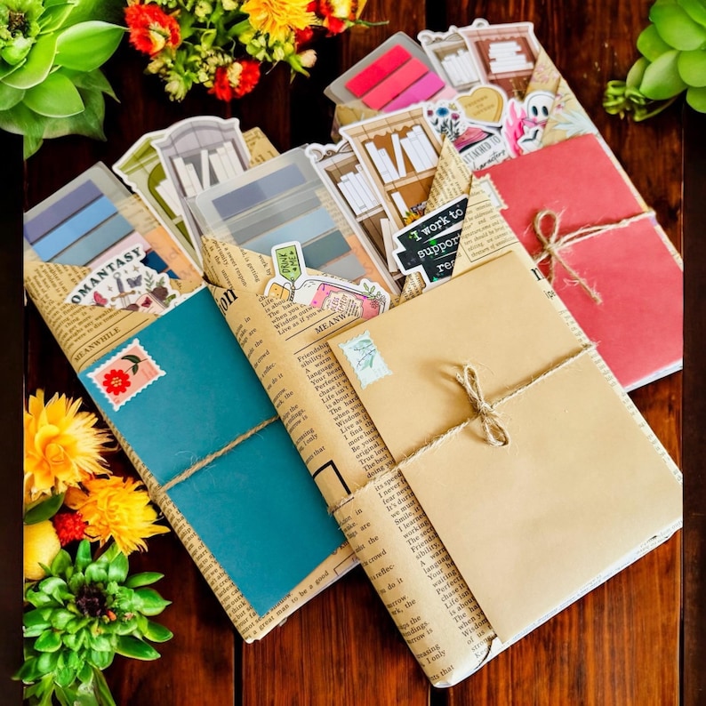 May include: A collection of handmade journals and notebooks. Each journal is wrapped in vintage newspaper and tied with twine. The journals feature colorful covers, stickers, and the text "OMANTASY" and "MEANWHILE".