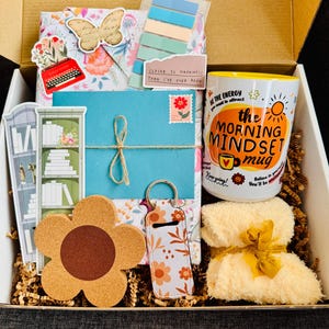May include: A gift box filled with various items, including a floral-patterned envelope tied with twine, a coffee mug with the text "The Morning Mindset Mug", a flower-shaped cork coaster, and a small towel tied with a yellow ribbon.