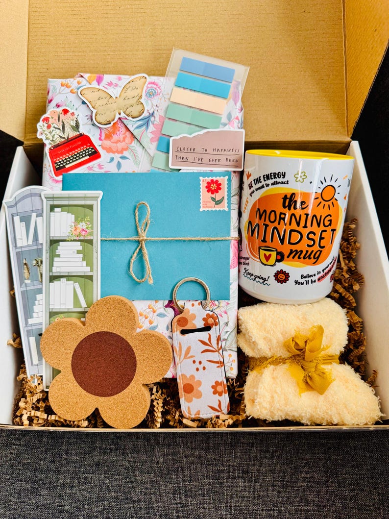 May include: A gift box filled with various items, including a floral-patterned envelope tied with twine, a coffee mug with the text "The Morning Mindset Mug", a flower-shaped cork coaster, and a small towel tied with a yellow ribbon.