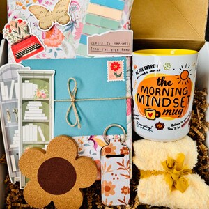 May include: A gift box filled with a variety of items, including a floral-themed mug with the text "the MORNING MINDSET mug", a flower-shaped coaster, a keychain, and a small yellow towel. Other items include a blue envelope, stickers, and decorative paper.