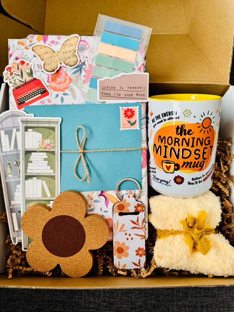 May include: A gift box filled with a variety of items, including a floral-themed mug with the text "the MORNING MINDSET mug", a flower-shaped coaster, a keychain, and a small yellow towel. Other items include a blue envelope, stickers, and decorative paper.
