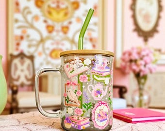 May include: A clear glass mug with a handle, a bamboo lid, and a green glass straw. The mug features a colorful graphic print of books, a teapot, a bird, and flowers, with the text "Pages of Peace" written on an open book illustration.