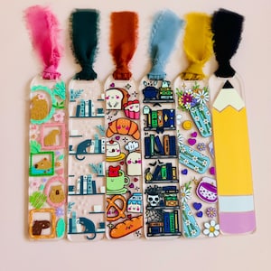 May include: Set of six clear acrylic bookmarks with colorful illustrations and ribbons. Designs include a capybara, cat, food, books, flowers, and a pencil. Each bookmark has a different colored ribbon attached at the top.