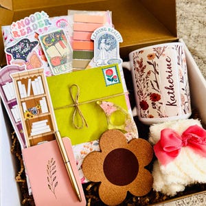 May include: A gift box filled with various items, including a floral-decorated mug with the name "Katherine", stickers, a pink notebook, a gold pen, a flower-shaped coaster, and a fluffy white item with a pink bow. The box contains a variety of stationery and accessories.