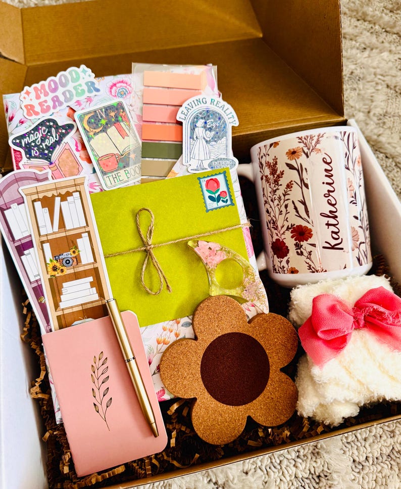 May include: A gift box filled with various items, including a floral-decorated mug with the name "Katherine", stickers, a pink notebook, a gold pen, a flower-shaped coaster, and a fluffy white item with a pink bow. The box contains a variety of stationery and accessories.