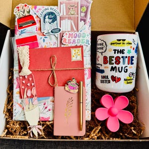 May include: A gift box filled with a variety of items. Includes a mug with the text "The Bestie Mug", a pink flower-shaped item, a pink notebook, a gold pen, and decorative stickers. The box is filled with shredded paper.