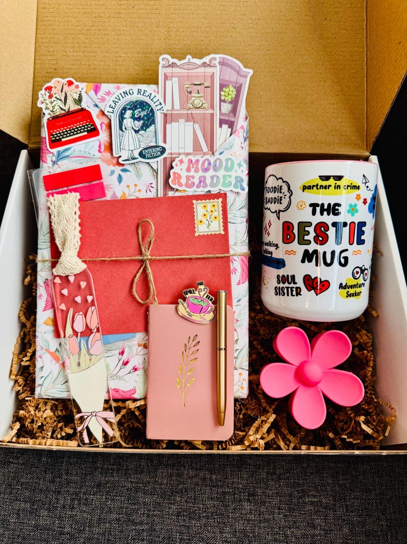 May include: A gift box filled with a variety of items. Includes a mug with the text "The Bestie Mug", a pink flower-shaped item, a pink notebook, a gold pen, and decorative stickers. The box is filled with shredded paper.