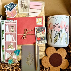 May include: A gift box filled with stationery and accessories. Includes a floral mug, stickers, a notebook, a pen, a coaster, and a red envelope tied with twine. The box contains a variety of items with a bookish or botanical theme.