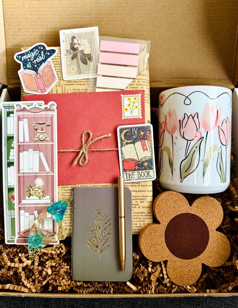 May include: A gift box filled with stationery and accessories. Includes a floral mug, stickers, a notebook, a pen, a coaster, and a red envelope tied with twine. The box contains a variety of items with a bookish or botanical theme.