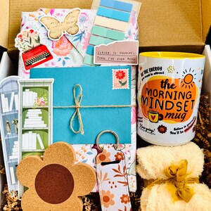 May include: A gift box filled with various items, including a floral-patterned package, a blue envelope tied with twine, a flower-shaped coaster, and a mug with the text "the MORNING MINDSET mug". The box also contains a keychain and a small towel.