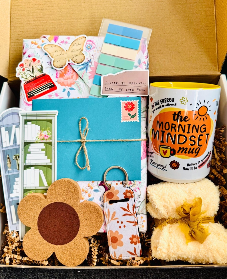 May include: A gift box filled with various items, including a floral-patterned package, a blue envelope tied with twine, a flower-shaped coaster, and a mug with the text "the MORNING MINDSET mug". The box also contains a keychain and a small towel.