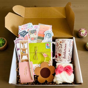 May include: A gift box filled with various items, including a floral-patterned mug with the name "Katherine", stickers, a pink notebook, a pen, a flower-shaped coaster, and a fluffy white sock with a pink bow. The box is open, revealing the contents.