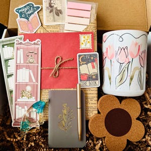 May include: A gift box filled with stationery and accessories. Includes a floral mug, a notebook with a gold floral design, a pen, stickers, a bookmark, a coaster, and a red envelope tied with twine. The box is filled with brown paper.