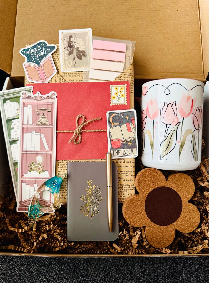 May include: A gift box filled with stationery and accessories. Includes a floral mug, a notebook with a gold floral design, a pen, stickers, a bookmark, a coaster, and a red envelope tied with twine. The box is filled with brown paper.