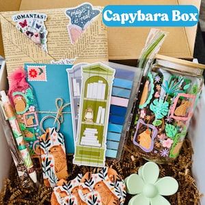 May include: A gift box labeled "Capybara Box" filled with various themed items. Includes stickers, a keychain, pens, coasters, a jar, and a flower-shaped clip. The items feature capybara illustrations and botanical designs, with a color palette of greens, oranges, and blues.