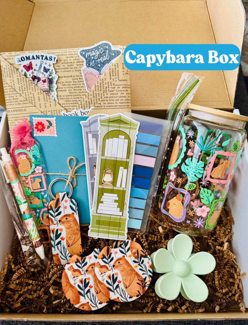 May include: A gift box labeled "Capybara Box" filled with various themed items. Includes stickers, a keychain, pens, coasters, a jar, and a flower-shaped clip. The items feature capybara illustrations and botanical designs, with a color palette of greens, oranges, and blues.