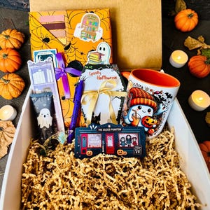 May include: A gift box filled with Halloween-themed items including a mug with a ghost illustration, a coffin-shaped notepad with a gold ribbon, a purple pen, a bookmark, a sticker, and a rectangular sign labeled The Gilded Phantom.