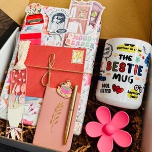 May include: A gift box filled with a white mug, a pink flower-shaped item, a pink notebook, a gold pen, and a red envelope tied with twine. The mug has the text "The Bestie Mug" and other graphics. The box contains decorative paper and a bookmark.