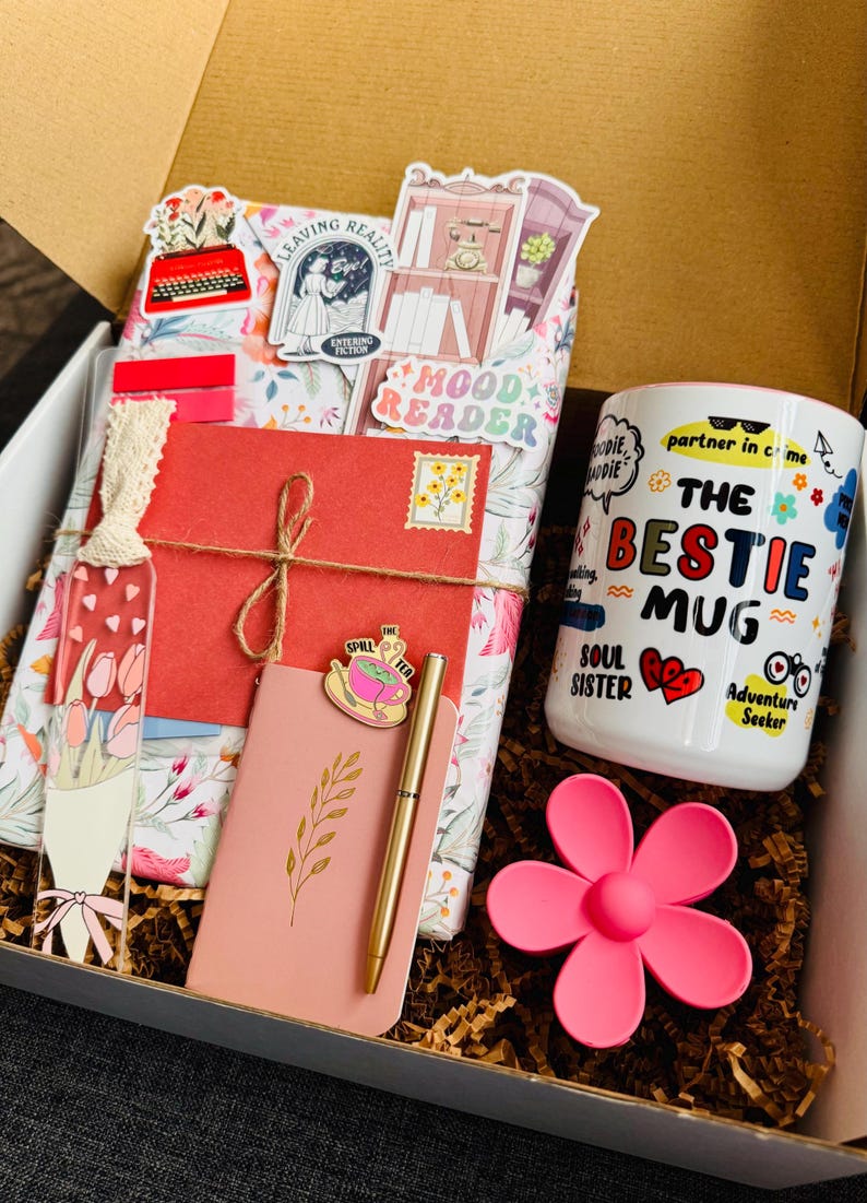 May include: A gift box filled with a white mug, a pink flower-shaped item, a pink notebook, a gold pen, and a red envelope tied with twine. The mug has the text "The Bestie Mug" and other graphics. The box contains decorative paper and a bookmark.