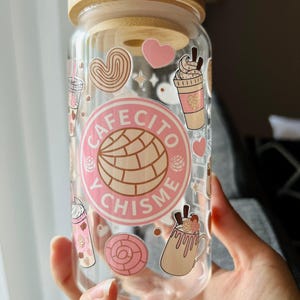 May include: A clear glass tumbler with a bamboo lid featuring pink and brown decals of concha sweet bread, coffee drinks, and hearts. The central circular graphic displays the text "CAFECITO Y CHISME" surrounding a concha illustration.