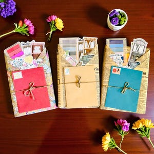 May include: Three decorative packages wrapped in newspaper print, each tied with twine and containing stationery items. The packages feature red, tan, and teal envelopes, adorned with stickers and stamps. Flowers and a small pot of purple flowers are arranged around the packages.