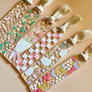 May include: A collection of clear acrylic bookmarks with various designs, including flowers, moons, and bows. Each bookmark has a cream-colored lace ribbon attached at the top. The designs feature pastel colors and intricate details, perfect for book lovers.