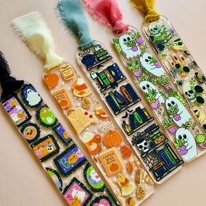 May include: A collection of Halloween-themed bookmarks with colorful ribbons. Each bookmark features unique designs, including ghosts, pumpkins, books, and the text "PUMPKIN SPICE RECIPES". The bookmarks are made of clear acrylic.