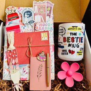 May include: A gift box filled with a variety of items. Includes a "The Bestie Mug", a pink notebook, a gold pen, a bookmark, and decorative stickers. The box is lined with brown paper and a pink flower.