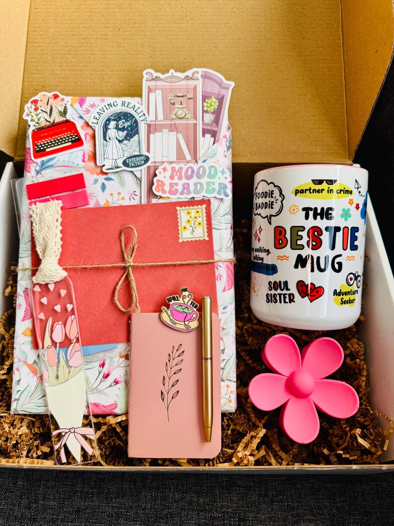May include: A gift box filled with a variety of items. Includes a "The Bestie Mug", a pink notebook, a gold pen, a bookmark, and decorative stickers. The box is lined with brown paper and a pink flower.