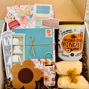 May include: A gift box filled with various items, including a floral-patterned journal, a coffee mug with the text "the MORNING MINDSET mug", a flower-shaped coaster, and a small, folded towel tied with a yellow ribbon. The box also contains stickers and a teal envelope.