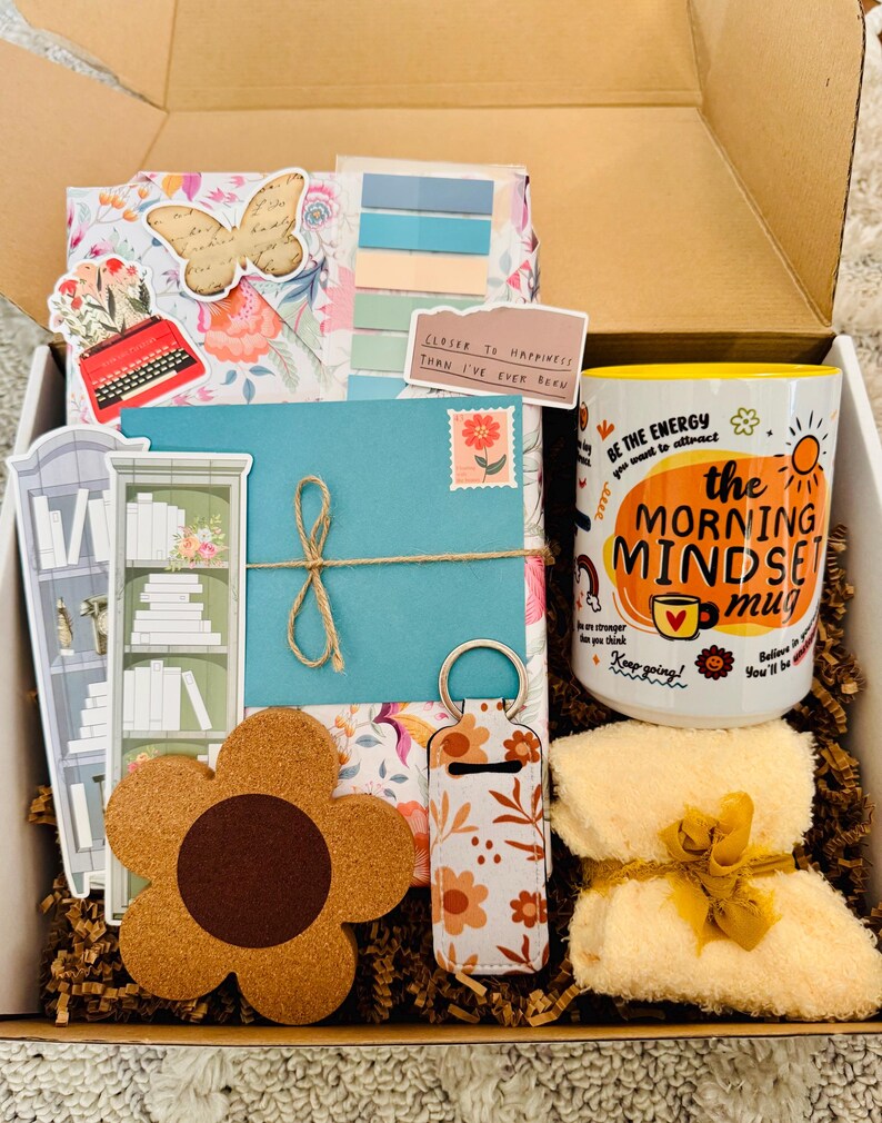 May include: A gift box filled with various items, including a floral-patterned journal, a coffee mug with the text "the MORNING MINDSET mug", a flower-shaped coaster, and a small, folded towel tied with a yellow ribbon. The box also contains stickers and a teal envelope.