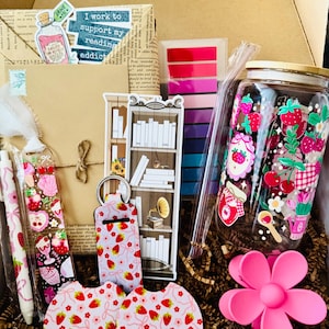 May include: A gift box filled with reading-themed items. Includes a jar with strawberry designs, stickers, pens, keychains, coasters, and a pink flower hair clip. The box also contains a bookmark with a bookshelf design and the text "I work to support my reading addict".