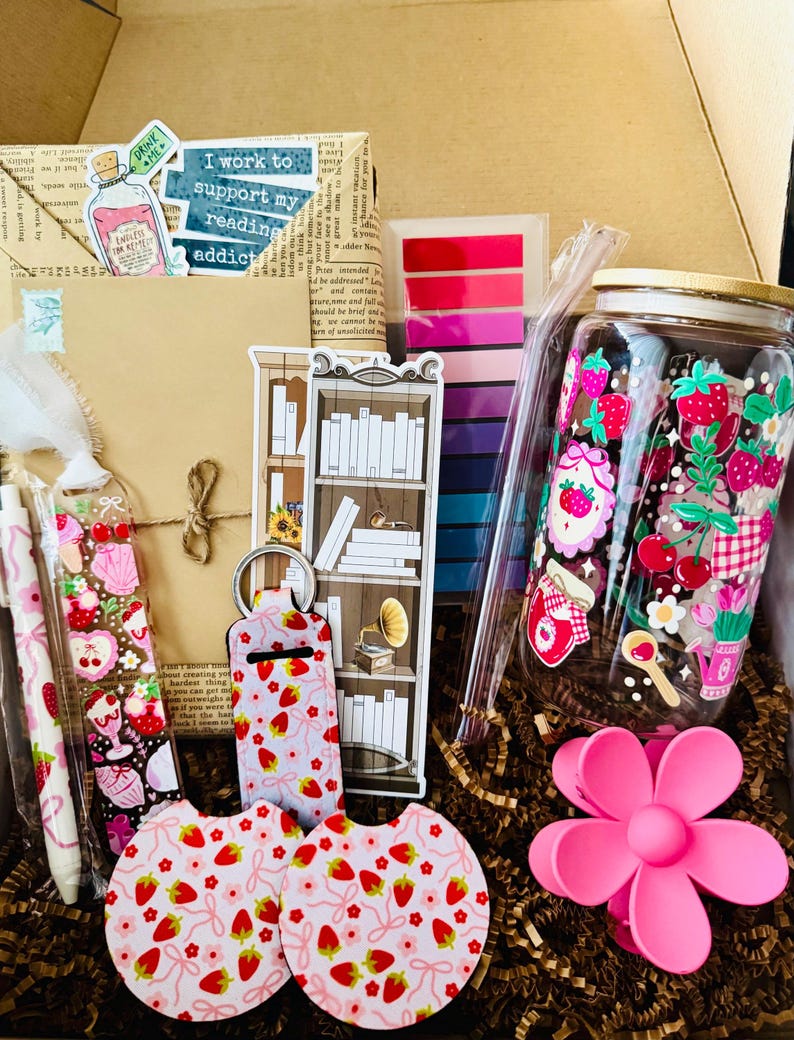 May include: A gift box filled with reading-themed items. Includes a jar with strawberry designs, stickers, pens, keychains, coasters, and a pink flower hair clip. The box also contains a bookmark with a bookshelf design and the text "I work to support my reading addict".