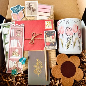 May include: An open gift box filled with stationery and accessories. Includes a floral mug, a notebook with gold floral design, a pen, stickers, bookmarks, a red envelope, and a flower-shaped cork coaster. The box is filled with shredded paper.