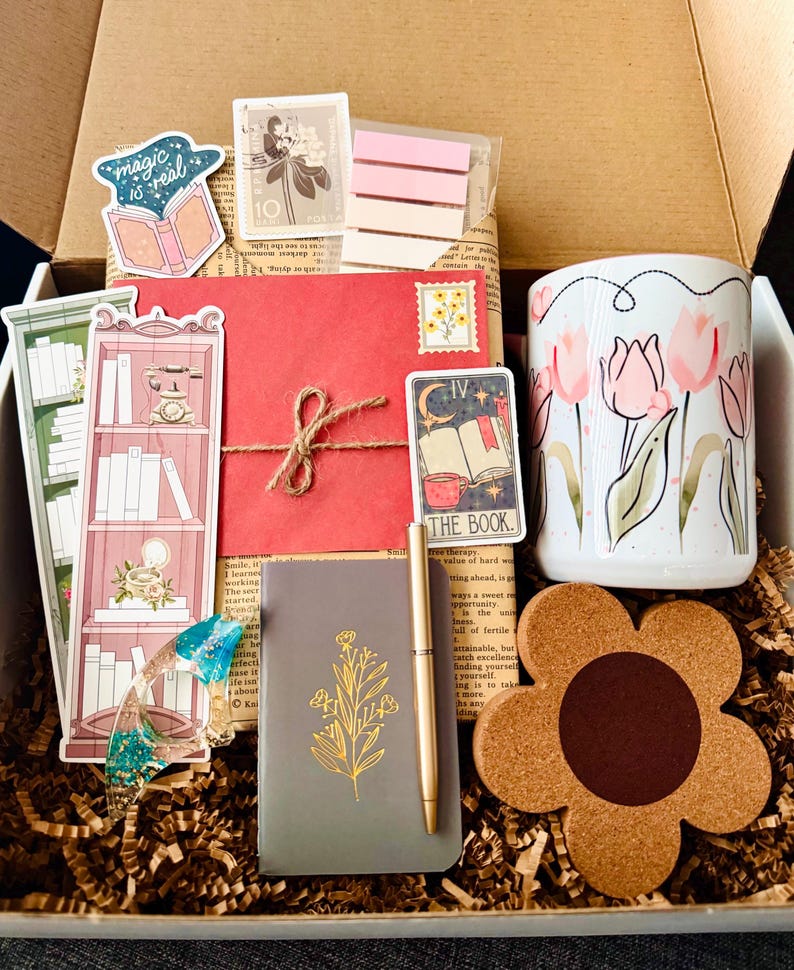 May include: An open gift box filled with stationery and accessories. Includes a floral mug, a notebook with gold floral design, a pen, stickers, bookmarks, a red envelope, and a flower-shaped cork coaster. The box is filled with shredded paper.