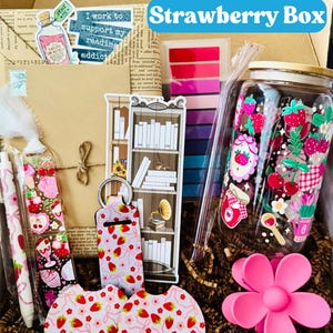 May include: A gift box labeled "Strawberry Box" filled with various items. Includes a decorated jar, pens, stickers, coasters, a keychain, and a flower-shaped phone holder. The items feature strawberry and floral designs.
