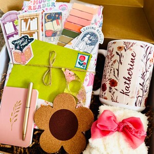 May include: Gift box filled with a variety of items. Includes a floral mug with the name "Katherine", a pink notebook and pen, a flower-shaped coaster, stickers, and a fluffy white item with a pink bow. The box is filled with brown crinkle paper.