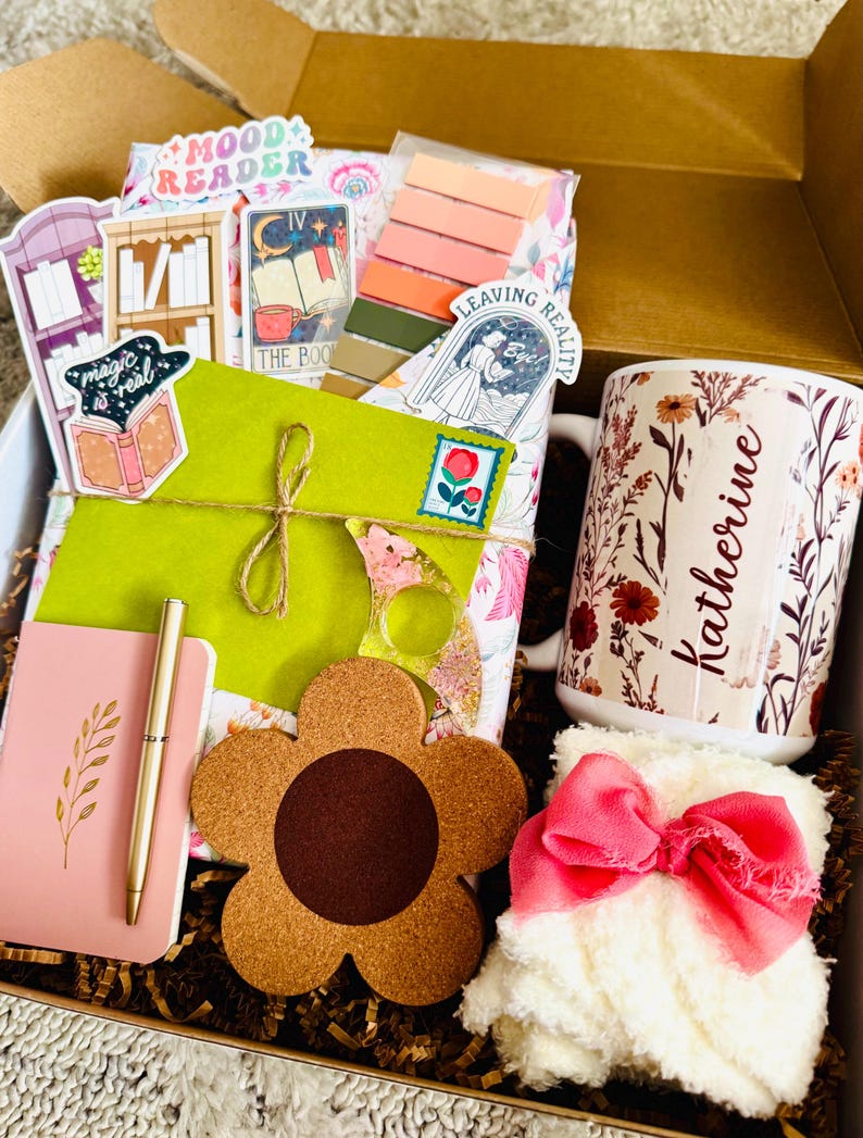 May include: Gift box filled with a variety of items. Includes a floral mug with the name "Katherine", a pink notebook and pen, a flower-shaped coaster, stickers, and a fluffy white item with a pink bow. The box is filled with brown crinkle paper.