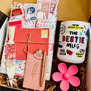 May include: A gift box filled with a mug, notebook, pen, and decorative items. The white mug has the text "THE BESTIE MUG" with colorful graphics. A pink flower-shaped item and a pink notebook with a gold pen are also visible.