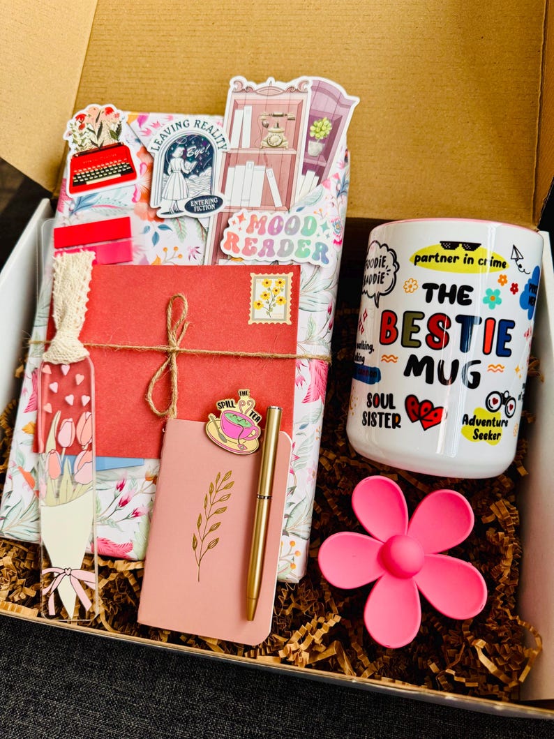 May include: A gift box filled with a mug, notebook, pen, and decorative items. The white mug has the text "THE BESTIE MUG" with colorful graphics. A pink flower-shaped item and a pink notebook with a gold pen are also visible.